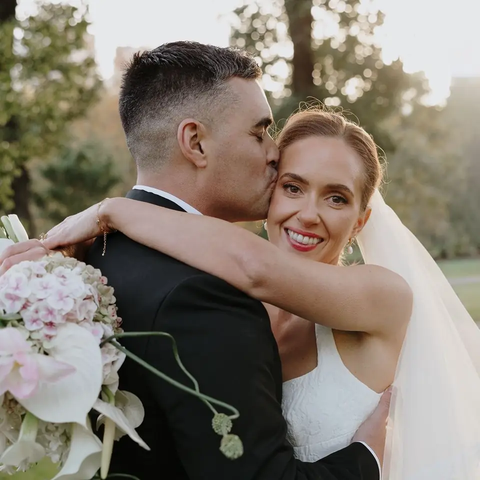 A bride smiling at the camera while her groom kisses her forehead on their wedding day.